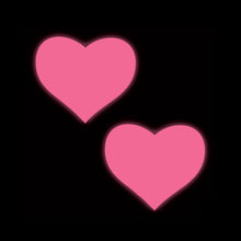 Load image into Gallery viewer, Two heart-shaped neon pink Pastease nipple covers glowing against a black background. These fun, flirty covers are designed to fit all breast shapes and sizes, handmade in the USA by a small family business. They're perfect for festivals, parties, and adding a cheeky touch to any outfit.