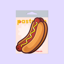 Load image into Gallery viewer, The Hotdog Pasties Weiner with Mustard Nipple Covers by Pastease®. Two hot dog sausages in long buns with mustard drizzled across in a zig zag pattern nipple covers shown in the pastease pastel packaging on a pastel purple background. Perfect for a festival, burlesque performance, drag shows, pride or parties.