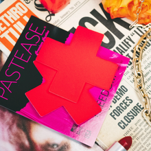 Load image into Gallery viewer, The Plus X: Neon Red (Blacklight Reactive) Cross Nipple Pasties by Pastease. Two neon Red Cross Plus X nipple covers in the pastease pink and black packaging surrounded by newspapers, lipstick and chain necklaces.