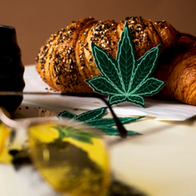 Load image into Gallery viewer, A pair of Indica Pot Leaf: Green Weed Nipple Pasties by Pastease leaning up against a croissant with a pair of sunglasses in a warm lit room.