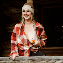 Load image into Gallery viewer, Model with hair in braids and wearing a bobble hat in a wooden cabin smiling and pulling plaid orange shirt to reveal her wearing the Pumpkin Spice Latte Breast Pasties Nipple Covers by Pastease, a glittery pumpkin spiced latte with whipped cream.