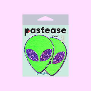 Alien: Neon Green/Glow in the Dark with Glittering Purple Eyes Nipple Pasties by Pastease®. Two green extraterrestrial heads with glitter eyes nipple covers shown in the pastease packaging on a pastel pink background.