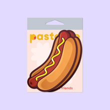 Load image into Gallery viewer, The Hotdog Pasties Weiner with Mustard Nipple Covers by Pastease®. Two hot dog sausages in long buns with mustard drizzled across in a zig zag pattern nipple covers shown in the pastease pastel packaging on a pastel purple background. Perfect for a festival, burlesque performance, drag shows, pride or parties.
