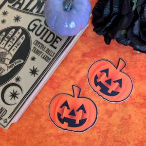 Pumpkin: Spooky Halloween Jack O' Lantern Nipple Pasties by Pastease. Two orange smiling carved pumpkins nipple covers on an orange fluffy background with various witchy items such as palmistry guides and black flowers surrounding them.