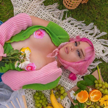 Load image into Gallery viewer, A femme model with pink hair wearing the Rose: Pink Glitter Velvet Blooming Rose Nipple Pasties by Pastease with an open green cardigan and pink wrist warmers on a picnic blanket outside.
