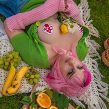 Load image into Gallery viewer, A pink haired alt femme model wearing the Shiny Red &amp; White Glow-in-the-Dark Shroom Nipple Pasties by Pastease with an open green cardigan with pink wrist warmers whilst laying down on a picnic blanket outside with fruit surrounding them.
