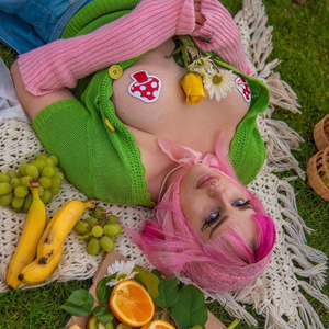 A pink haired alt femme model wearing the Shiny Red & White Glow-in-the-Dark Shroom Nipple Pasties by Pastease with an open green cardigan with pink wrist warmers whilst laying down on a picnic blanket outside with fruit surrounding them.