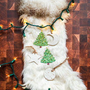 Christmas Tree Nipple Pasties by Pastease®. Two green glittery Christmas tree shaped nipple covers, shown on white fur background with star and moon Christmas decorations and fairylights. Perfect for a festival, burlesque performance, pride, Christmas or parties