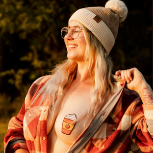 Model in plaid orange shirt and bobble hat smiling in the golden sun on an Autumn's day with shirt pulled to the side to reveal the Pastease pasties Pumpkin Spices nipple cover nipple cover on model's breast.