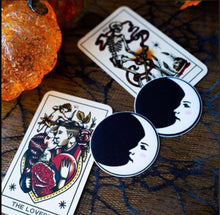 Load image into Gallery viewer, Two Moon: Black and White Man in the Moon Nipple Pasties by Pastease shown next to two tarot cards and glass pumpkins on a black net table cover.
